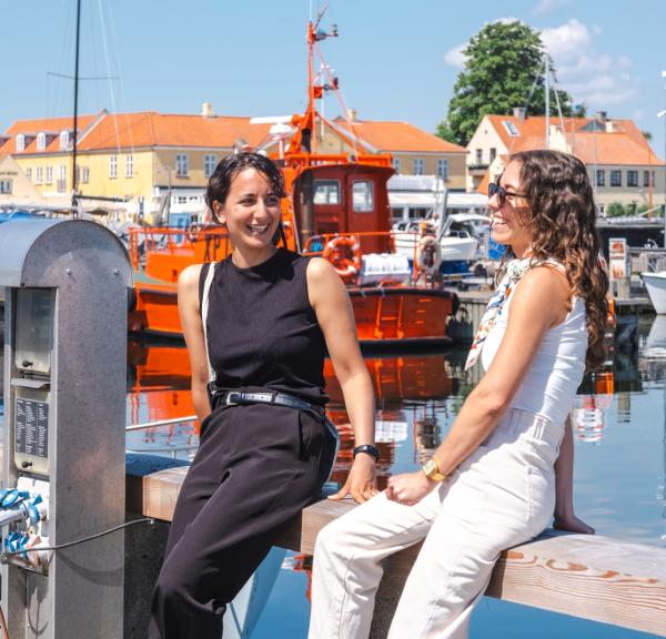 To kvinder sidder på rækværk på havnen
